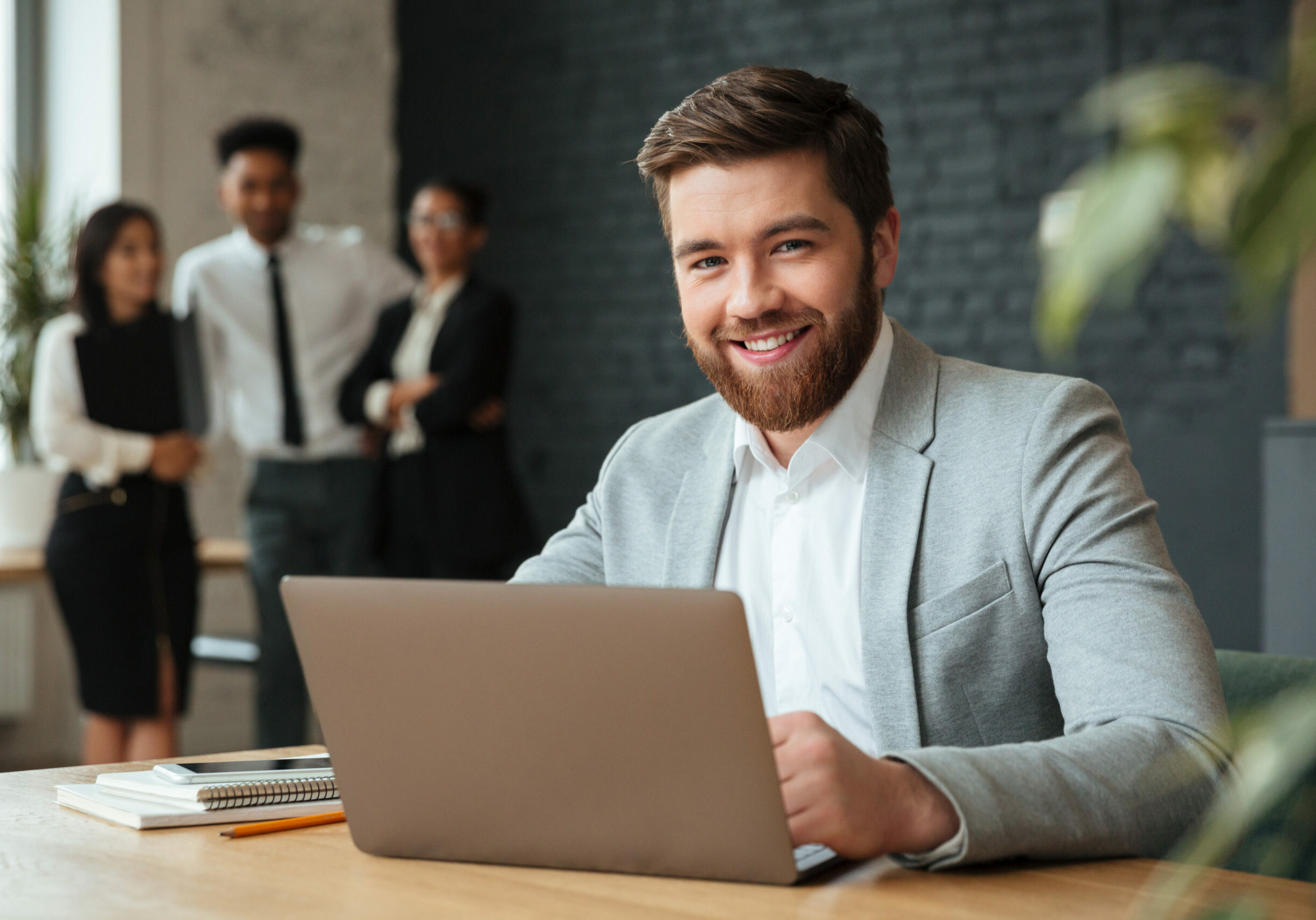 Image of cheerful young caucasian businessman sitting indoors using laptop computer. Looking camera.